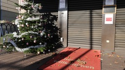 Ein Weihnachtsbaum steht neben einem heruntergelassenen Rolladen in einer Fußgängerpassage. Bild: picture alliance / augenklick/Ralf Ibing/firo Sport © picture alliance / augenklick/Ralf Ibing/firo Sport | Ralf Ibing Foto: Ralf Ibing