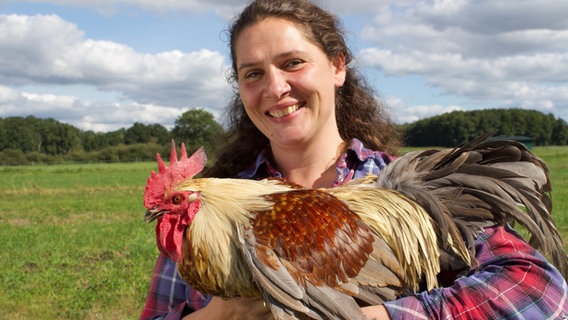 Geflügelbäuerin Christine Bremer auf ihrem Hof in Kölau (Niedersachsen) aus den "Hofgeschichten - Ackern zwischen Alpen und Ostsee" mit einem Hahn auf dem Arm. © NDR/Hoheluft Filmproduktion Foto: Peter Janßen