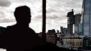 Ein ehemaliger Merrill-Lynch-Banker sitzt verschattet (unkenntlich gemacht) vor der Kulisse der Londoner Skyline in einem Hotelzimmer. © NDR/ARD 