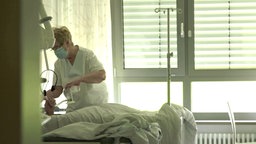 Eine Person mit Munschutz und Kittel beugt sich zu einer Person, die im Krankenbett liegt © NDR Foto: Screenshot