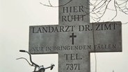 Grabkreuz mit der ironischen Inschrift: Hier ruht Landarzt Dr. Zimt. Nur in dringenden Fällen, Tel: 7371  