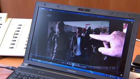 Eine Hand zeigt auf ein Videobild, das auf einem Laptop zu sehen ist.  