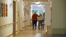 Eine Pflegerin läuft mit einer Seniorin durch einen Pflegeheimflur und legt dabei den Arm um sie. © NDR 