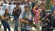 Ein Mann steht mit einem Maschinengewehr vor einer Demonstration © NDR/ARD Foto: NDR/ARD